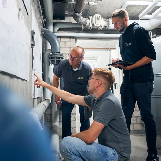 Betreiber, Vertreter von Gebr. KEMPER und der CAD-Agentur Lehmann stehen im Technikraum vor einem Revisionsplan. Der Betreiber erläutert anhand des Planes die bestehende Anlage, während von den Vertretern Fragen und Notizen auf einem Tablet notiert werden. 