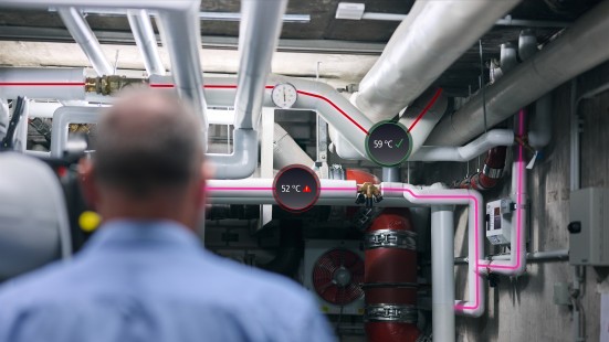 Haustechniker steht im Technikraum vor einer komplexen, schwer überschaubaren Trinkwasserinstallation. Die Rohrkonstruktion wird digital angedeutet, einige ablesbare Temperaturen liegen außerhalb des Sollbereiches.